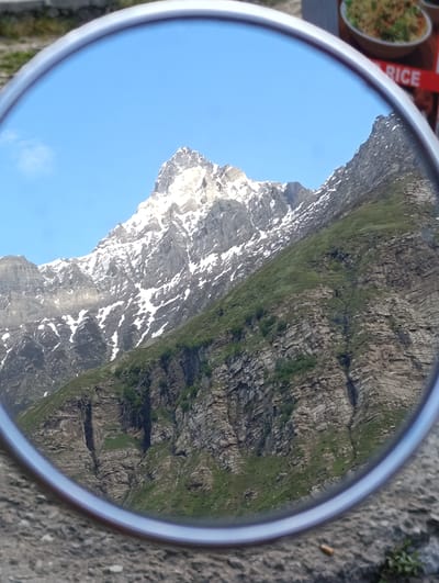 The image displays a reflection in a round mirror. The reflection shows a majestic mountain range with snow-capped peaks and green slopes. The sky is a clear blue. The mirror is placed on a rough, gray surface, possibly a stone path or ground. In the background, slightly out of focus, a red sign with white text that says "RICE" is partially visible, along with a bowl of food, suggesting a roadside eatery or a stall. The mountain scenery appears to be in a remote, high-altitude location in India.