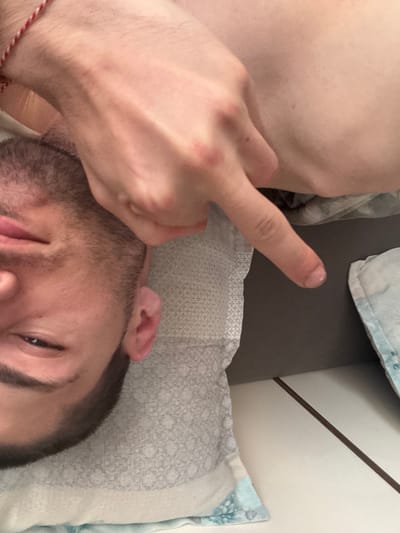 A man lies on a patterned pillow, his face turned towards the camera. He has short, dark hair and a stubbled beard. His right hand is raised, index finger pointing downwards towards what appears to be a grey surface and a corner of bedding. A red and white string bracelet is visible on his wrist. The image is close-up, focusing on the man's face and hand.