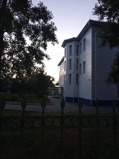 The image depicts a modern, multi-story building with a white tiled facade, accented with a dark blue base. The building appears to be a school or administrative complex, situated in a grassy area with trees. A dark green metal fence with decorative circular elements runs along the foreground, partially obscuring the view of the grounds. Behind the fence, a paved walkway leads towards the building, which has several visible windows and a prominent corner section. The time of day appears to be late afternoon or early evening, as indicated by the soft, diffused light and the warm glow of the setting sun filtering through the trees. The sky is a clear, pale blue, suggesting fair weather. The scene conveys a sense of calm and order, with the modern architecture contrasting with the natural elements of the trees and grass. There is no visible text in the image. The location is indicated as Yurga, Russia.