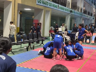 The image shows an indoor sports facility in Phagwara, India, likely a gymnasium or court for sports such as kabaddi, evidenced by the mats on the floor and the presence of individuals in athletic attire. A banner at the top reads "GO HARD GO HOME WINNER NEVER STOP." Several young women are present, some in blue athletic uniforms, and others in casual clothing. Some are sitting on chairs in the background, while others are gathered on the floor, seemingly engaged in a sports activity or discussion. A few individuals are in focus in the foreground, with one woman wearing a dark blue shirt with the word "REGION" visible on the back. The environment includes seating, sports equipment like water bottles, and safety netting around the playing area. The lighting appears to be artificial, typical of an indoor sports venue.