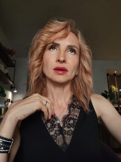A woman with shoulder-length, wavy, blonde hair is shown from the chest up. She is wearing a black sleeveless top with a lace neckline. A silver necklace with a pendant is visible. She has red lipstick on and her left hand is resting on her chest, with a ring on her right index finger. The background is dimly lit and appears to be an indoor setting. There are shelves with objects on the left side of the image, and a lamp is visible. The image does not contain information about the city of Faliukovo, Russia.