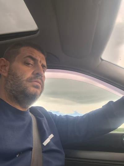 A man with a beard is seen from the chest up, inside a car. He is wearing a dark blue sweatshirt with a white rectangular patch on the left breast. A grey seatbelt is visible across his chest. The man is looking towards the camera with a serious expression. The interior of the car, including the sunroof and window frame, is visible. The outdoor environment appears cloudy with some hints of blue sky. The image was taken in Dolno Sahrane, Bulgaria.