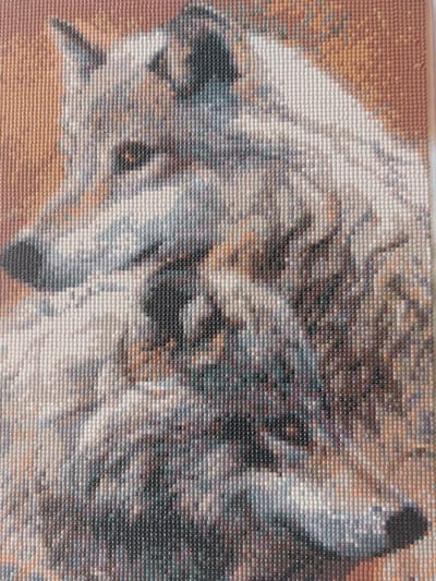 The image displays a close-up view of a diamond painting, likely a wolf's head and upper body, rendered in a mosaic of small, colored resin beads. The painting is characterized by its pixelated appearance, with individual beads forming the intricate details of the wolf's fur, eyes, and snout. The color palette ranges from warm earthy tones for the background to cooler grays, whites, and blacks for the wolf itself, with subtle hints of purple and blue in the shadows. The texture of the beads is evident, creating a shimmering, faceted surface. The image focuses on the artistic representation of the animal, with no discernible urban or environmental context. The painting does not depict any specific location within the city of Sad, Ukraine, as it is a standalone artwork.