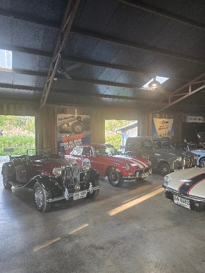 The image displays the interior of a garage or car display facility located in Ban Sahakon 2, Thailand. The structure features a dark, insulated ceiling with exposed metal truss work, a visible ceiling fan, a skylight, and a suspended light fixture. Dark wood-paneled walls are adorned with vintage automobile posters, including one depicting a race car with the text "MOTORING NEWS GRAND PRIX" and another for "SPORTS & SPRINGS." A light grey concrete floor, marked with scuffs and shadows, exhibits bright streaks of natural sunlight entering from openings to the left and right of the frame. Five classic vehicles are parked on the concrete surface. In the foreground left is a dark red and black vintage convertible roadster with a prominent chrome grille, round headlights, wire-spoke wheels, and a Thai license plate reading "กง 1556." Behind it, a red convertible sports car with white racing stripes is positioned. Further back on the right, a grey vintage sedan is parked, and a light blue vintage car is partially visible behind it. The front section of a white vehicle with blue racing stripes and a Thai license plate "ภค 8982" is visible in the foreground right. No individuals are present, and the scene is static.