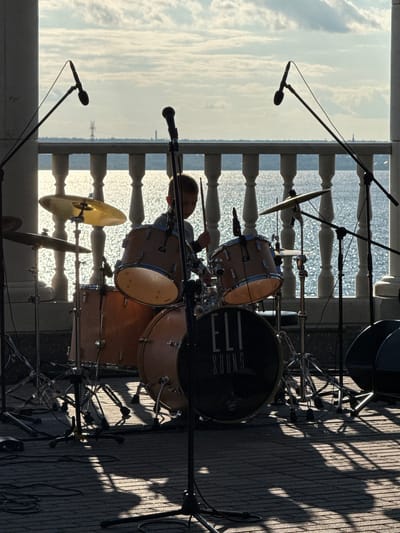 In the city of Sredny Postol, Russia, an outdoor stage is set up with a drum kit and microphones. A young person, silhouetted against the bright sky and shimmering water, is playing the drums. The drum kit includes multiple drums, cymbals, and stands, all of which are partially obscured by shadows cast by the surrounding architecture. The scene is lit by the setting sun, creating dramatic light and shadow patterns on the stage floor. A white balustrade separates the stage area from the water, and a few distant structures are visible on the horizon. The atmosphere suggests a performance or practice session taking place in a scenic outdoor location.