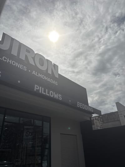 The image shows the exterior of a store in San Rafael, Costa Rica, located at latitude 9.938, longitude -84.153.  The store is named "Jiron" and sells pillows, bedding, and almohadas (pillows in Spanish). The store is located on a street corner and has a large, black sign with white lettering. The sign is partially obscured by the sun, which is shining brightly in the sky. The sky is mostly cloudy, but there are some patches of blue. The store has a large glass window and a metal roll-up door. The building is painted white. To the right of the store is a high, black wall, and behind that wall is a building with a grey roof.  There is a power line running horizontally across the top of the image, with a single white wire.  There are no people visible in the image.