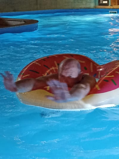 An adult male is seen in a swimming pool, floating on an inflatable donut. The man is bald and appears to be of Caucasian descent. He is wearing what seems to be a swimming suit, although it is not fully visible. His arms are outstretched, and his hands are blurred due to motion. The inflatable donut is red with yellow and green sprinkles, resembling a frosted pastry. The water in the pool is bright blue and appears slightly rippled. In the background, a portion of the pool's edge is visible, with a darker blue trim. The overall environment suggests an indoor or covered swimming area, as indicated by the dimly lit, flat ceiling in the upper portion of the image. The image was taken in the city of Sorbulak, Kazakhstan.