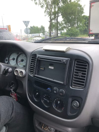 The image shows the interior of a car's dashboard and windshield, with a view of the outdoors through the rain-streaked glass. The dashboard features a radio or navigation unit, air vents, climate controls, a steering wheel with a key in the ignition, and gauges for RPM and speed. Outside, trees, a road, and a truck are visible. The scene appears to be in an outdoor urban or semi-urban environment, possibly Safonovo, Russia, given the context. The weather is overcast and rainy, as indicated by the water droplets on the windshield and the general lighting. There are no people clearly visible within the car.