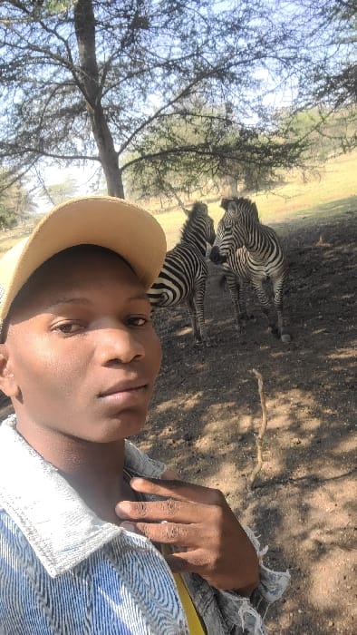 The image portrays an outdoor scene in the Mto wa Mbu area of Tanzania, featuring a person and two zebras. In the left foreground, an individual with dark skin wears a light-colored baseball cap and a light blue and white patterned shirt, with a yellow garment visible underneath. Their right hand rests near their collarbone. The person looks directly at the camera, indicating a self-portrait. In the midground, to the right of the person, two zebras with characteristic black and white stripes stand on dry, light brown ground. One zebra is positioned slightly behind the other. The background shows sparse trees with thin branches and minimal foliage, under a bright, clear sky. The terrain behind the zebras gently slopes upwards, exhibiting additional sparse vegetation, consistent with a savanna environment.