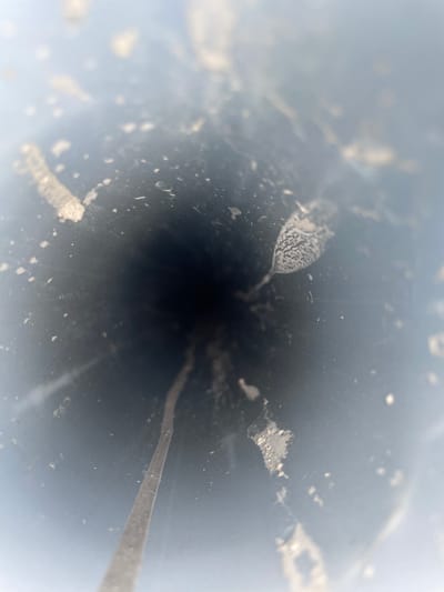 The image displays an internal, longitudinal view looking into a dark, cylindrical pipe or conduit, located in Mns Asan, Indonesia. The interior surface is dark gray to black, heavily speckled with numerous light-colored particulate matter, and exhibits irregular streaks and patches consistent with dust, residue, or mineral deposits. A prominent, thin, light-colored streak runs from the bottom-center foreground into the dark central depth of the conduit, following its curvature. On the right interior wall, a distinct, approximately 1.5 cm long, leaf-shaped or tear-drop-shaped patch with a finely textured or granular pattern is visible. The outer rim of the conduit, at the periphery of the image, appears lighter and out of focus, indicating ambient light entry and a shallow depth of field. No human subjects, specific actions, or external environmental elements are discernible within the frame.