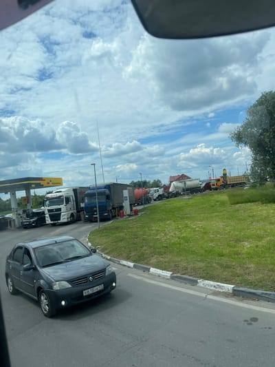 The image shows a queue of trucks and a car on a paved road next to a grassy area. The scene appears to be a roadside, possibly near a gas station, given the presence of a yellow and black sign that says "РАСПРОДАЖА". The sky is filled with white, fluffy clouds against a blue backdrop, suggesting daytime with fair weather. The trucks are of various types, including what appear to be cement mixers and flatbed trucks. In the foreground, a dark-colored car is visible with a license plate that reads "У 618 НК 162". The orientation of the image is rotated, making it appear as if the road is vertical and the sky is to the left. The overall impression is of a normal day with traffic on a road in what is suggested to be Старое Бобренево, Russia.