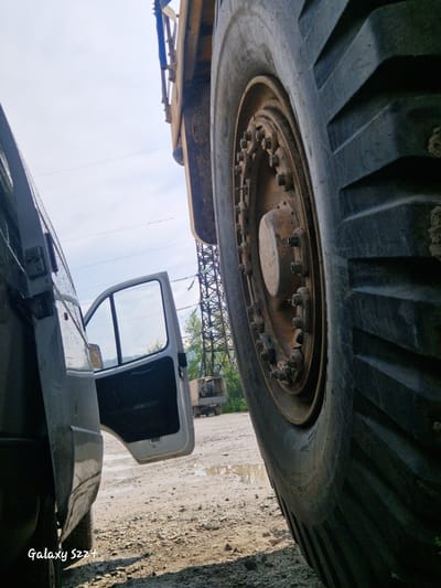 A low-angle, medium shot shows the immense tire of a heavy-duty vehicle dominating the foreground, its treads deeply grooved and covered in dirt. To the left, a white van is parked, its driver's side door ajar, revealing a dark interior. Beyond the van, the ground is a mix of dirt and puddles, indicative of recent rain. In the background, a tall power pylon stands against a hazy sky, with trees and another vehicle visible further in the distance. The scene appears to be an outdoor industrial or construction area, possibly on the outskirts of Zheleznogorsk-Ilimsky, Russia. The "Galaxy S22+" watermark is visible in the bottom left corner of the image.