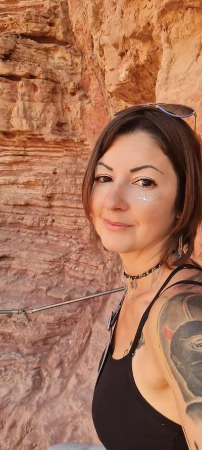 A woman with shoulder-length brown hair stands in front of a geological formation of orange and brown rock. She wears a black tank top and has multiple tattoos visible on her chest and shoulder. Sunglasses are propped on her head. She has glitter makeup around her eyes and a nose piercing. She wears a beaded necklace with metal chains hanging from it. The background is composed of the striated rock formation. The location is likely in the Hevel Eilot Regional Council, Israel, due to the rock formation resembling those in the region.