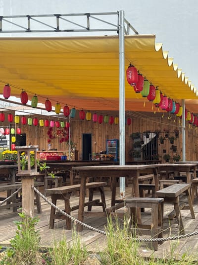 This image depicts an unoccupied outdoor or semi-outdoor seating area of a commercial establishment in Hòa Cường Ward, Vietnam. A large yellow retractable awning, supported by a silver metal truss structure, covers the scene from above. Strung beneath the awning are multiple lines of various colored cylindrical paper lanterns, predominantly red, green, yellow, pink, and blue, each featuring white floral patterns.

The ground consists of a weathered wooden deck or platform, partially edged by green grass and small plants in the immediate foreground. A thick, braided rope serves as a low barrier or railing on the wooden deck. Numerous rustic wooden tables and matching benches are arranged across the deck, facing a wooden-paneled building or wall that forms the background. The furniture appears robust and aged.

Against the wooden background, a counter or serving area is visible, displaying various items and adorned with red decorative elements, possibly artificial flowers or small lanterns. A blue Pepsi-branded upright refrigerator or cooler stands near this counter area. To the left, a display of yellow potted flowers, likely chrysanthemums, is visible on a table. On the right side of the background wall, several small potted plants are hung vertically. Two dark doorways are visible within the wooden wall structure. No individuals are present in the scene, and no actions or interactions are depicted.