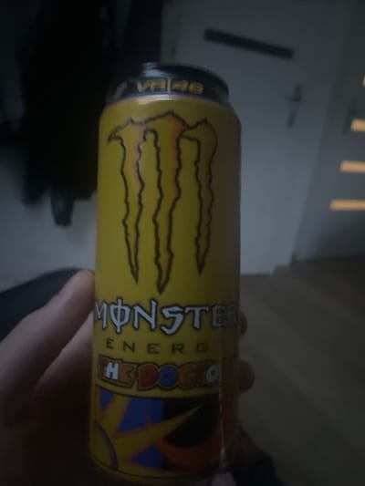 In Paczkowo, Poland, a person's hand is holding a yellow can of Monster Energy drink. The can features the iconic Monster logo, the "VR46" designation, and the text "THE DOGO." The background is dark and out of focus, suggesting an indoor setting with dim lighting.