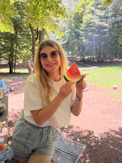 The image depicts an outdoor gathering in a wooded area within Dolovi, Montenegro, under bright daylight. A blonde female with sunglasses and a white t-shirt stands centrally, smiling at the viewer while holding a triangular slice of red watermelon with black seeds. She wears light blue denim shorts and a dark bracelet on her left wrist. To her left, partially visible on a picnic table, are two large blue water bottles, a small silver pot, and a dark speckled mug. A blue plastic bag containing another watermelon slice rests near the table. In the mid-ground on the left, a male figure wearing a grey t-shirt and dark trousers is visible from the back, facing away from the camera. The ground in the foreground and mid-ground consists of reddish-brown dirt with scattered dry leaves and twigs. A metal barbecue grill rack lies on the ground in the bottom right foreground. Beyond the dirt path, a bright green grassy area extends into the background, where a deflated or dirty white soccer ball is visible on the grass and several rough logs or benches are arranged. The scene is heavily treed, with dense green foliage creating dappled sunlight and shadows on the ground.