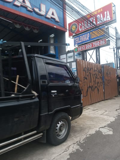 The image shows a black pickup truck parked on the side of a street in front of a business. The business has a sign that reads "CAHAYA BAJA" and "TERLENGKAP TER". There is a phone number on the sign. There is also a corrugated metal fence with graffiti on it. Above the fence are signs for Spyro, Taso Kaso Metal, Cahaya Baja, and Bluescope. The location is likely in Asia, given the text on the signs.
