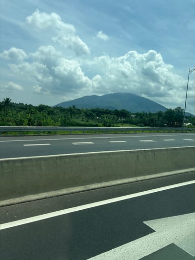 The image depicts a highway in Binh Xuan 2, Vietnam, under a cloudy sky. In the foreground, a concrete barrier runs along the side of the road, separating it from the highway lanes marked with white lines. Beyond the highway, a lush green landscape unfolds, featuring dense vegetation and banana trees. In the background, a large, verdant mountain rises, its slopes covered in trees and foliage. A single lamppost is visible on the right side of the image. The overall scene is one of natural beauty juxtaposed with man-made infrastructure.