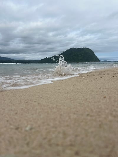 A low-angle, eye-level shot captures a wave splashing onto a sandy beach in Babahie, Indonesia. The foreground is dominated by the textured sand, with some small pebbles visible. In the mid-ground, the ocean is a tranquil blue, with a frothy white wave breaking and sending water droplets into the air. Beyond the ocean, a lush green island rises majestically, its slopes covered in dense vegetation. The sky above is overcast with dramatic, cloudy formations. The overall scene evokes a sense of natural beauty and the power of the ocean.