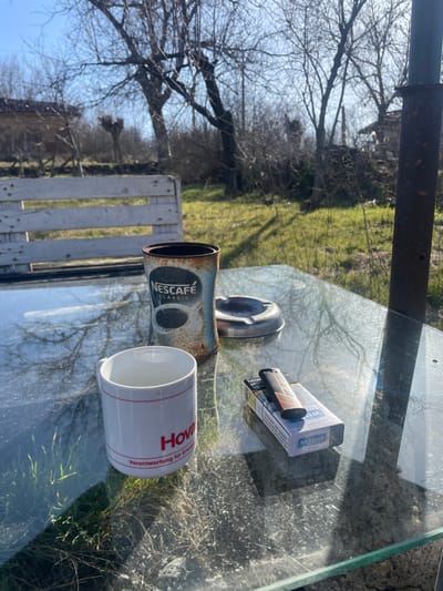 An outdoor scene in Topchii, Bulgaria, captures a glass-topped table under direct sunlight. On the table, from left to right, are a white ceramic mug with "Hoval Verantwortung für Energie" printed in red, a rusty metal Nescafe Classic can, a silver-colored metal ashtray, a pack of Karelia 100s cigarettes, and a black and orange lighter resting atop the cigarette pack. The table surface is dusty. In the background, a grassy area with patches of dry vegetation is visible. Several bare deciduous trees, indicative of late winter or early spring, dominate the midground. To the left, a white wooden bench or pallet structure is present. A rusty metal pole is partially visible on the far right. The sky is clear blue. No individuals are present.