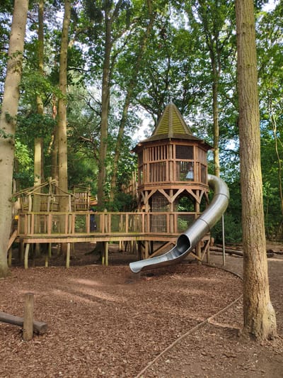The image depicts a wooden adventure playground structure within a forest setting. The playground features a multi-level wooden platform with railings, a circular tower with a conical roof, and a long, winding metal slide. A young person wearing a blue shirt and dark shorts is standing on the lower level of the platform, looking to the left. Another young person, wearing a light-colored dress, is visible inside the circular tower. The ground beneath the playground is covered in wood chips. The surrounding environment is densely populated with tall trees, their canopies creating a dappled light effect on the ground. The image appears to be taken in the daytime, with bright, natural light filtering through the trees. The style of the playground suggests it is designed for children's recreation. It is likely located in a park or woodland area. While the image itself does not explicitly identify the location, such playgrounds are common in family-friendly outdoor spaces. Without further information, a specific location within the city of Rugby, United Kingdom cannot be determined.