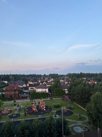 The image captures a panoramic view of a suburban residential area, likely in Военнослужащих Кп, Russia, during twilight. The sky exhibits a gradient of soft blue and pink hues, with a visible full moon high in the frame. Below the sky, a dense canopy of lush green trees stretches across the horizon, interspersed with the rooftops of numerous houses. The architecture of the houses appears to be of a modern design, with varied roof colors and window placements.
In the foreground, a well-maintained children's playground is prominently featured. The playground consists of multiple brightly colored play structures, including slides, swings, and climbing equipment, all situated on a soft, dark rubberized surface. Surrounding the playground are manicured green lawns, winding paved pathways, and decorative landscaping. A small, decorative pond with aquatic plants is visible in the lower right corner, bordered by a wooden fence. The overall scene suggests a peaceful and family-friendly environment. There are no visible people in the image.