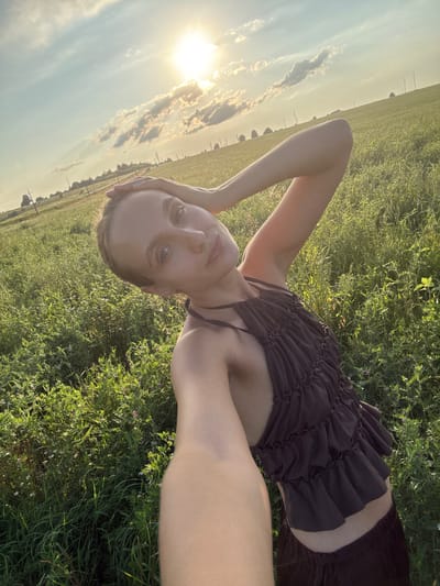 The image shows a young person lying on their back in a grassy field. The sun is setting, casting a warm glow over the scene. The person is wearing a dark, ruffled top and dark shorts. Their left arm is bent, with their hand resting on their head, and their right arm is extended. The person's face is tilted towards the camera, and they appear to be looking at the viewer with a relaxed expression. The background shows a vast field with some trees and power lines in the distance, indicating a rural setting. The sky is a mix of blue and clouds, with the sun's rays breaking through. The overall mood of the image is peaceful and serene, capturing a moment of quiet reflection in nature. There is no visible text in the image.