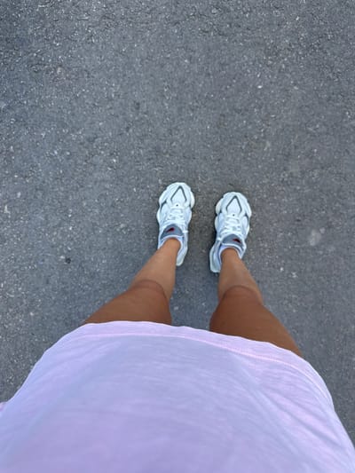 The image depicts a person's legs and feet from a top-down perspective, standing on an asphalt surface. The individual is wearing white sneakers with gray and red accents, and a light pink t-shirt is visible around the waist. The asphalt appears to be a city street or sidewalk, likely in Serpneve, Ukraine, given the context provided. The lighting suggests it is daytime. There are no other individuals or objects in the immediate vicinity.