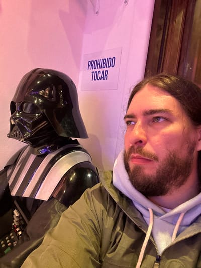 An indoor scene, rotated 90 degrees clockwise, features a man posing with a Darth Vader display under pinkish ambient lighting.
In the upper portion of the frame (when viewed upright), a man is visible from the chest up. He has long brown hair and a beard, and his gaze is directed downwards and to the left. He is dressed in a light blue hooded sweatshirt underneath a green jacket, which features prominent pink drawstrings.
Below the man, the upper torso and head of a Darth Vader display or mannequin are prominently featured. The iconic black helmet with its characteristic mask is clearly visible, along with the upper chest plate adorned with black and silver striped details.
The background consists of a plain white wall illuminated with a soft pinkish-purple hue. Attached to this wall is a white sign with blue lettering that reads "PROHIBIDO TOCAR," which translates to "FORBIDDEN TO TOUCH." In the very top of the frame, a dark wooden structure, possibly part of a door frame or ceiling, is partially visible.
The image captures a moment, likely in a themed exhibition, museum, or retail space in Villa General Belgrano, Argentina, given the visible text and context. The man appears to be observing or interacting with the display, possibly taking a photo, with the specific lighting contributing to a distinctive atmosphere.