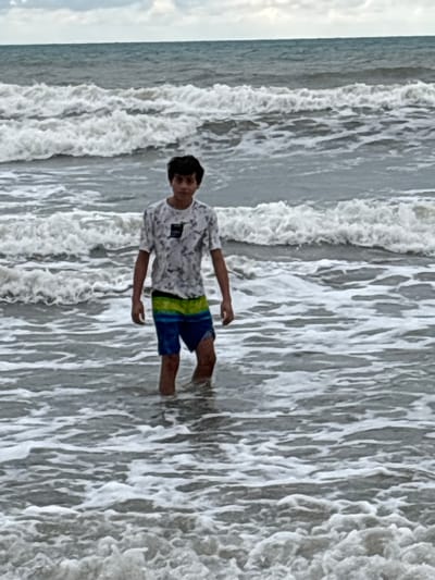 A person is standing in the ocean. The waves are crashing around them, and their clothes are getting wet. The person is wearing a patterned short sleeve shirt and colorful blue and green swim trunks. The ocean is a dark color, and the sky is overcast. The waves are foamy and white.