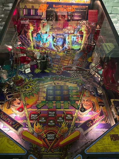 This image captures the illuminated playfield of a "Ripley's Believe It or Not!" pinball machine, located in Winterthur, Switzerland. The machine is powered on, with various lights and displays glowing, indicating it is ready for play.
The playfield is richly detailed with a tropical adventure theme, featuring cartoon-style depictions of Robert Ripley and a monkey-like character. Graphics include palm trees, bamboo, and ancient-looking idols. Prominent elements on the playfield include multiple ramps, targets, and bumpers. Two yellow-and-red flippers are visible at the bottom of the playfield, with their corresponding "Believe It" and "or Not!" labels.
Notable game features and text are visible across the machine. At the top left, a red sign shows "Scramble Temple Code" with options "B. Wake Up Idol" and "C. Amazing Event". Other text includes "TOMB STONE", "IDOLS COLLECTED" with numbers 1, 2, 3, "SKILL", "BOZO", and multiple "SPECIAL" indicators. The right side of the playfield highlights score opportunities such as "ROAD TRIP", "BIG POINTS", "LITE EXTRA BALL", and "SUPER JACKPOT VALUE" ranging from "2 MILLION" to "6 MILLION", alongside "LITE SUPER JACKPOT" and "SHOOT AGAIN". Reflections are visible on the clear glass cover, caused by the machine's own lights and ambient room lighting, suggesting an indoor setting during evening or night. The artwork at the very bottom right mentions "DESIGNED & MANUFACTURED IN THE U.S.A. BY THE PINDALL PEOPLE IN THE WORLD" and "PRESS FLIPPERS." The machine appears well-maintained and vibrant.