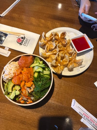 A wooden table holds a bowl of sushi with salmon, avocado, seaweed, cucumber, and rice. Beside the bowl are chopsticks on a napkin and a plate of fried wontons with a red dipping sauce. A hand holds a cell phone near the wontons. Packets of branded materials rest to the side.