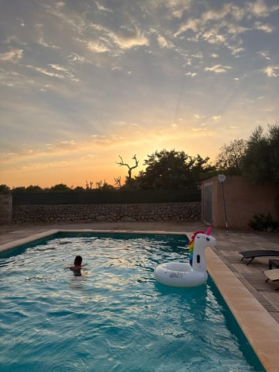 In Santa Margalida, Spain, at sunset, a person swims in a rectangular swimming pool. An inflatable unicorn float is partially submerged in the pool near the edge, which is lined with beige coping and a wooden deck. A pool skimmer on a pole leans against a building in the background. The sky displays a warm gradient of orange and yellow, with scattered clouds and sunbeams radiating downwards. The property is bordered by a dry stone wall and a dark fence, with trees and vegetation visible beyond.