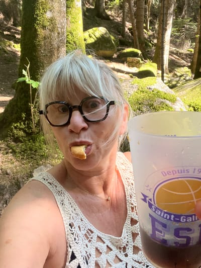 An outdoor selfie shows a woman in Chazelles-sur-Lavieu, France, in a forest setting with moss-covered rocks and trees. The woman, who appears to be middle-aged, has blonde hair, is wearing glasses, and is holding a drink in a translucent plastic cup. She is wearing a cream-colored, crocheted top over a dark garment. In her mouth, she has a piece of bread or pastry. The background consists of trees, foliage, and dappled sunlight.