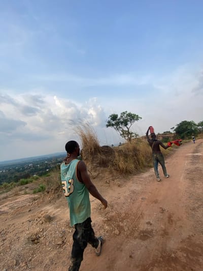 The image depicts an outdoor scene in Asaga Amangwu, Nigeria, on a rugged, uphill dirt path. In the foreground, a dark-complexioned male, seen from behind, walks towards the left, looking downhill. He wears a faded light-green basketball jersey with "LYELL 33" on the back, dark, possibly mud-splattered pants, and light-colored sandals. His right hand is raised to the back of his neck. Further up the dirt path, in the midground, another dark-complexioned male walks away from the camera. He is shirtless, wearing blue jeans and light-colored shoes. He holds a red bucket or container overhead with his right hand and a bundle of red fabric in his left hand. A small, distant figure is visible further up the path. The environment consists of dry, reddish-brown soil, scattered rocks, patches of tall, dry grass, and sparse green trees. A valley with a distant settlement and green vegetation is visible on the left side, under a partly cloudy sky with light blue and white clouds.