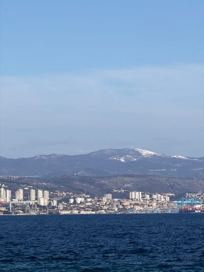 The image shows a view of Grad Opatija, Croatia from the sea. A mountain range runs parallel to the coast with some snow visible. A city is situated between the mountains and the sea. A port can be seen at the northern end of the city. The water is dark blue with visible ripples. The sky is light blue, getting paler towards the horizon.