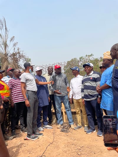 A group of approximately a dozen men is gathered outdoors on a bright, clear day in Aloko, Nigeria. They stand on reddish-brown dirt ground, facing forward and slightly to the right of the frame. Most are dressed in casual attire, including various shirts, trousers, and baseball caps.
The central activity involves a man in a plaid shirt who is holding a microphone and gesturing with his left hand, appearing to be speaking or addressing the group. Other men surround him, some looking towards him with attentive expressions, while a few smile. One individual, positioned behind the main speaker on the far right, is holding a smartphone and seemingly recording the event.
In the background, a structure made of metal reinforcing bars (rebar) is visible, suggesting a construction site or an area undergoing development. Sparse, dry-looking vegetation can also be seen. The bright, cloudless sky indicates clear weather during daytime. While some of the caps worn by the men have logos or text, none are clearly legible.