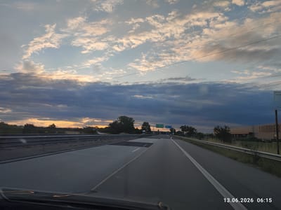 The image shows a view from a car traveling on a highway. The sky is filled with dramatic clouds, with the sun setting in the distance on the left side of the frame, casting a warm glow. The highway stretches ahead, with lane markings visible. There are trees and vegetation on the sides of the road, and in the distance, a building with "LIEBHERR" signage is visible. Traffic signs are also present, indicating directions to destinations such as "Benkovski" and "Plovdiv". The timestamp "13.06.2026 06:15" is present in the lower right corner. The scene appears to be located on a highway leading towards or near Benkovski, Bulgaria, based on the visible signage.