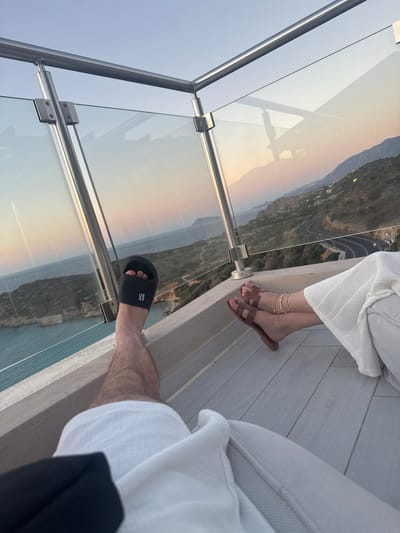 Two people are on a balcony with a glass railing overlooking a coastal landscape. One person, wearing white clothing, has their legs crossed and is wearing brown slides and ankle bracelets. The other person, also wearing white clothing, is wearing a black slide. The scene appears to be during sunset, with warm colors reflecting on the glass railing. The location appears to be a resort or a scenic overlook, given the view of the sea and the mountainous terrain. There is a road visible winding through the landscape. The overall mood seems relaxed and peaceful, consistent with a vacation or a leisurely outing. The text "BIO ALL" is visible on the black slide.