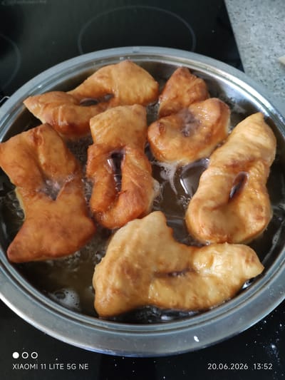 This is a close-up, overhead, eye-level shot of several golden-brown, irregularly shaped pieces of fried dough, likely pastries, submerged in hot oil within a silver metal frying pan. The pastries have a slightly puffed and uneven texture, with some appearing to have been twisted or folded before frying, creating indentations or "holes" in their center. Bubbles and ripples from the hot oil are visible around the pastries, indicating they are actively cooking. The pan is situated on a dark, flat stovetop, possibly an induction cooktop, with some faint reflections visible. The background is blurred, but appears to be a kitchen environment. A date and time stamp "20.06.2026 13:52" and text "XIAOMI 11 LITE 5G NE" are present in the lower right and left corners respectively.

While the image depicts food being prepared, there is no information within the image itself that indicates a specific location in the city of Böblingen, Germany.