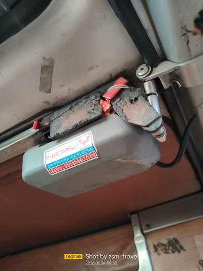 This is a close-up, slightly low-angle, outdoor photograph of a gray plastic electrical enclosure mounted on a metal frame, likely part of an automated gate mechanism. The enclosure is dusty and shows some wear and tear. A sticker on the enclosure reads "PHUONG CHUYEN CUA LUAA TU DONG Zalo: 0906661694", which translates to "PHUONG AUTOMATIC DOOR SPECIALIST Zalo: 0906661694". The background consists of a light-colored metal surface and a darker, possibly wooden surface below. The scene appears to be located in a general urban or semi-urban environment.