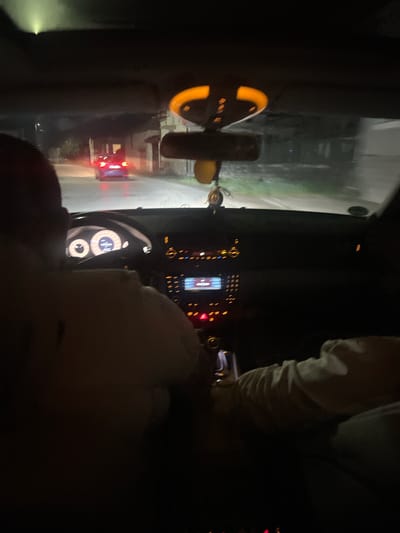 The image shows the interior of a car at night. The dashboard and steering wheel are visible, as well as the driver and front passenger, partially obscured by darkness. The exterior view through the windshield shows a road illuminated by headlights, with buildings and other vehicles. There is no information in the photo to locate the place in Gorna Rositsa, Bulgaria.