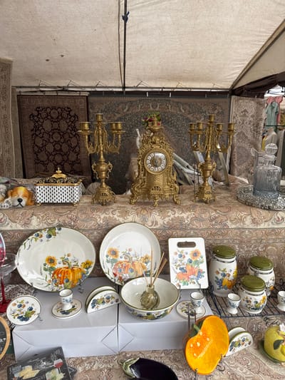 The image shows a close-up view of a market stall in посёлок 1-го отделения совхоза "Масловский", Russia. The stall is set up under a canvas awning, displaying various home decor items and tableware. Dominating the foreground are ornate brass candelabras and a gilded mantel clock. The table is covered with a richly patterned tapestry, on which are arranged ceramic dishes decorated with floral and pumpkin motifs, as well as a bright orange ceramic pumpkin-shaped bowl. Other items include glass decanters, a small decorative box, and what appear to be stacks of white paper bags. The lighting suggests it could be daytime, with no specific weather conditions discernible. The overall impression is of a vendor displaying a collection of decorative items, possibly for sale at a flea market or craft fair. There is no visible text in the image.