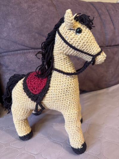 A crocheted toy horse, light tan with black mane, tail, and hooves, sits on a textured purple sofa. The horse has a red saddle with a black trim, and a black halter. The background shows the textured purple upholstery of the sofa. The scene is indoors and there are no people visible. This image is not located in Proletarsky, Russia.