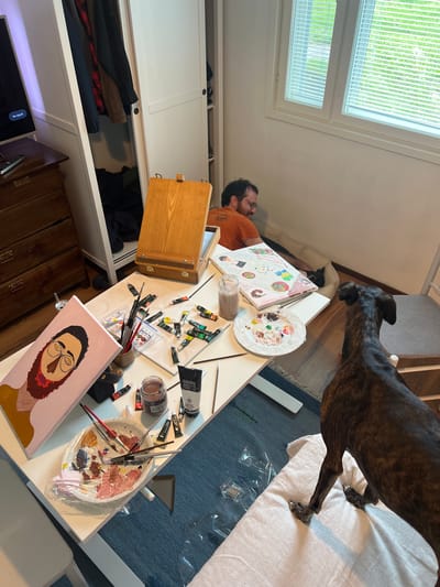 In a room with white walls and a wooden floor, a man in an orange t-shirt is lying on a dog bed, looking at a painting on a table. The table is covered in art supplies, including paint tubes, brushes, and a palette. A canvas with a portrait of a person is leaning against a wooden easel. A brindle-colored dog stands near the table, looking at the man. A closet with clothes is visible in the background. The room appears to be a living space in Hämeenlinna, Finland, possibly an apartment or house.