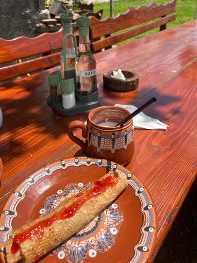 On a sunny day in Meshtitsa, Bulgaria, a wooden picnic table holds a Bulgarian-themed breakfast. A terracotta plate with a blue and white patterned rim holds a rolled crepe topped with red jam. A matching terracotta mug with a handle contains milk and a dark straw. Next to the mug is a small wooden ring holding a crumpled white tissue. Further down the table sits a grey stand containing three glass bottles labeled "ОЛІО," with olive-green dispensers. The table itself is made of reddish-brown stained wood, with a matching attached bench visible in the background.