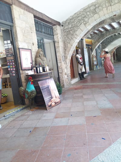 The image depicts an outdoor passageway in Besalú, Spain, characterized by stone architecture featuring arches. In the foreground, a colorful, decorative rooster statue stands to the left. Behind it, a wooden barrel serves as a display for bottles of wine or spirits. A framed picture rests on top of the barrel, and a printed advertisement lies on the ground next to it. Further down the passageway, a man dressed as a knight stands guard. To the right, a woman in a red and white patterned dress walks away from the viewer, adjusting her white hat. The ground is paved with terracotta-colored tiles.