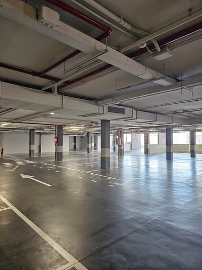 This media file depicts the interior of a parking garage in Cuarte de Huerva, Spain. The scene is captured from a low angle, looking upwards towards the ceiling and the expansive, empty concrete floor marked with white lines indicating parking spaces and directional arrows. The perspective emphasizes the repetitive structure of the garage, with rows of concrete pillars and overhead ductwork visible.
The lighting suggests daytime, with sunlight streaming in from openings in the garage, casting long shadows across the floor and creating a sense of depth. The overall atmosphere is utilitarian and quiet, with no people or vehicles present, highlighting the emptiness of the space.
Notable details include various pipes, conduits, and electrical boxes mounted along the wall and ceiling, indicative of the garage's infrastructure. Some pipes are painted red, possibly for fire suppression systems. The concrete surfaces show signs of wear, typical of a functional parking facility. The focus is on the architectural elements and the stark geometry of the parking garage. There is no visible text in the image, apart from the directional arrows and lane markings on the floor.