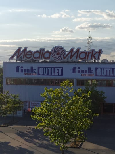 The image captures the exterior of a large retail building in Bischofsheim, Germany, featuring prominent signage for "Media Markt" and "fink OUTLET SCHUHE." The Media Markt sign is a large red and white illuminated logo with a swirl design. Below this, a banner displays "fink OUTLET SCHUHE" in purple and red lettering. The building itself is a light-colored, multi-story structure with large windows. The surrounding environment includes trees with lush green foliage casting shadows on the asphalt ground, suggesting it is daytime with sunlight. In the background, a tall electricity pylon is visible against a sky with scattered clouds. A smaller sign on the building's facade reads "Schuhe reduziert," indicating a shoe sale. There are no people visible in the image. The scene appears to be a commercial district.