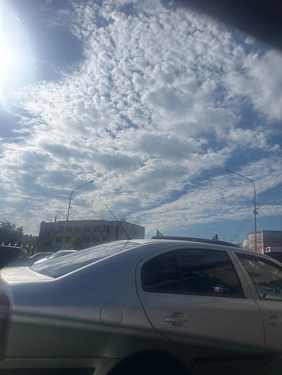 The image shows a silver sedan parked on a street in Ruzhyn, Ukraine. The sky is filled with white, fluffy clouds, and the sun is shining brightly, casting a lens flare in the upper left corner. In the background, there is a multi-story building with many windows and a street lamp. The car is viewed from the side, with its rear end and passenger side doors visible. The windows of the car are tinted, and some reflections can be seen on the surface of the vehicle. Other cars and trees are also partially visible in the background, suggesting an urban or suburban setting.
